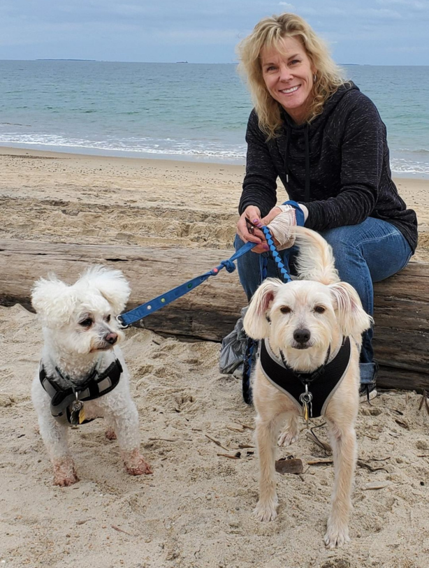 Woman sitting on a beach log holding two small dogs on leashes by the shoreline